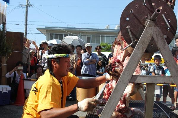 ねじり鉢巻きに黄色い法被姿の男性が牛の丸焼きを包丁でカットしようとしている写真